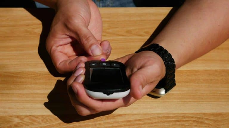 A person doing an at-home finger-prick blood test with a compact glucose and ketone meter on a wooden table.